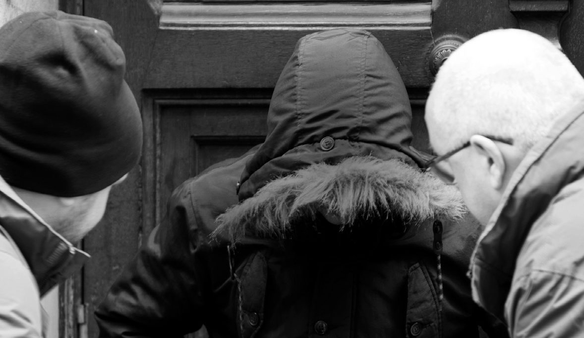Black and white image of man in beanie hat, man in black hooded coat looking down and man with glasses - two men looking at hooded man