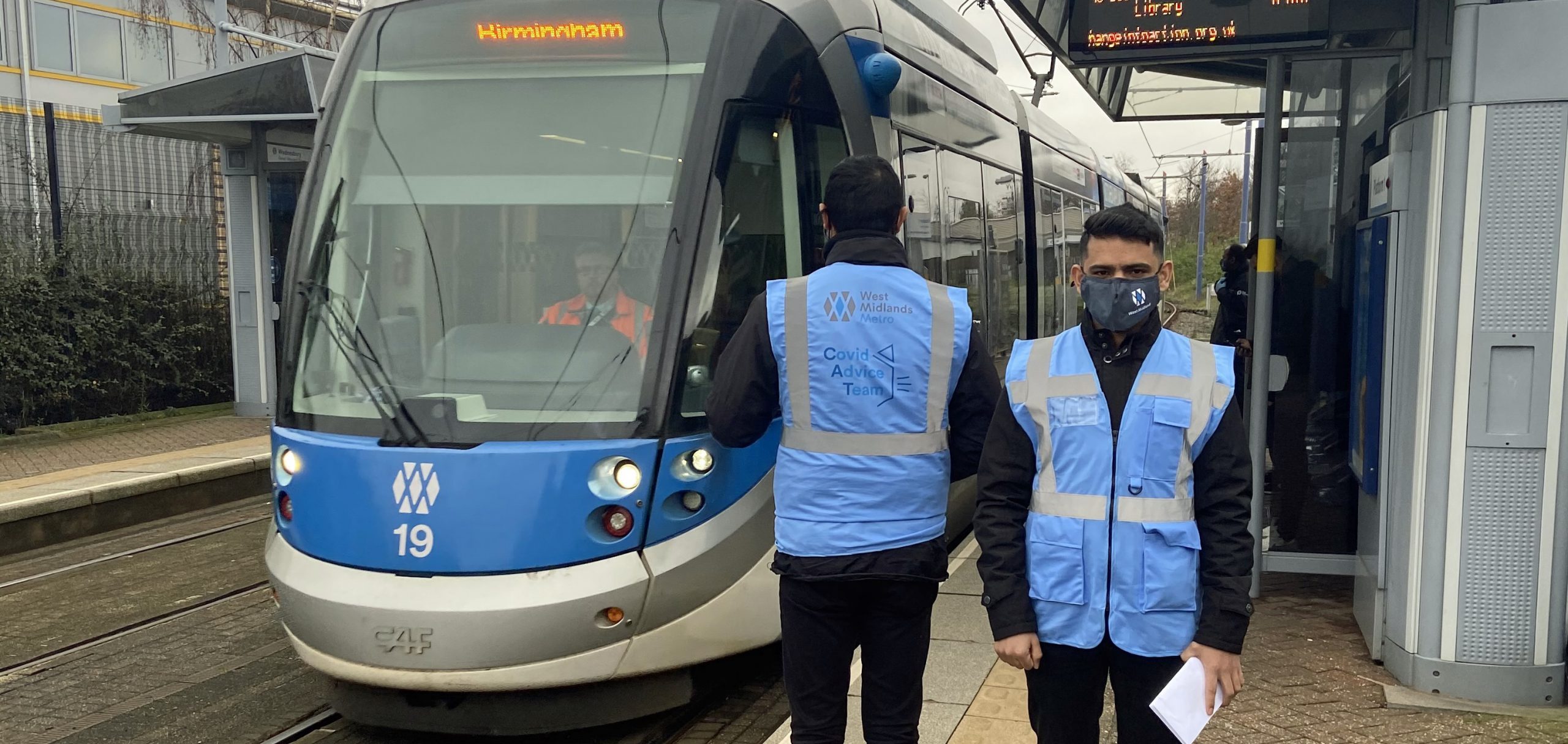 Image of tram with two men in high vis standing on platform next to it - One man is facing the tram, one is facing the camera wearing a face mask