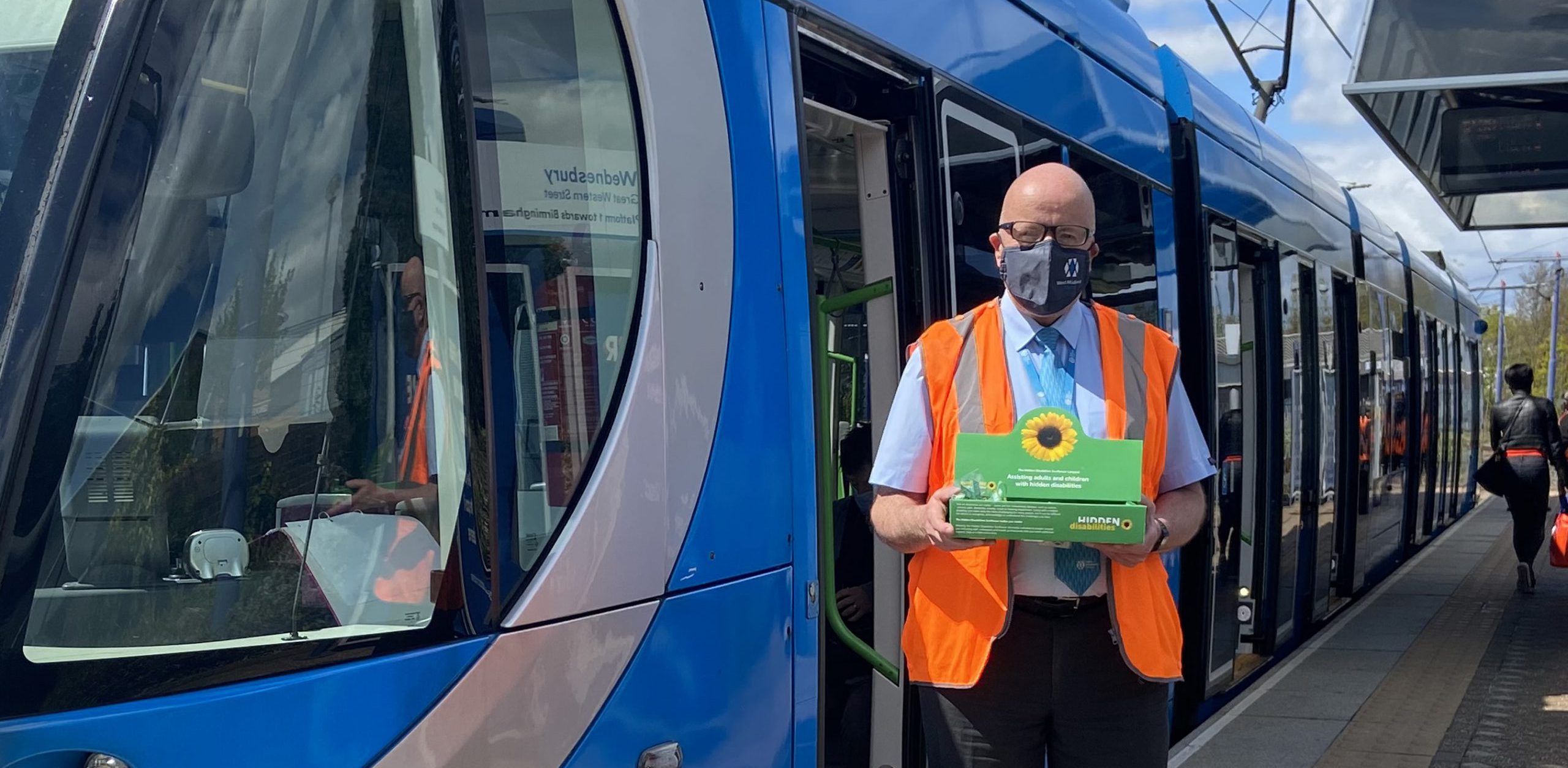 Man in front of tram wearing high vis and face mask holding sunflower charity donation box